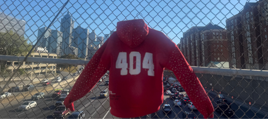 Red hoodie with '404' printed on it hanging against a chain-link fence with a cityscape in the background.