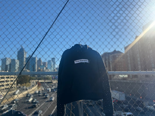 Black jacket with a brand logo against a cityscape with a chain-link fence.
